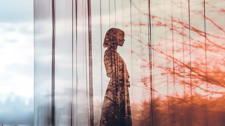 Double exposure of a silhouette of a woman standing behind sheer curtains and branches of a tree against a soft