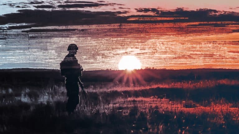 Double exposure of a silhouette of a soldier standing in a field and a vibrant red and orange sunset: solitude and resilience concept.